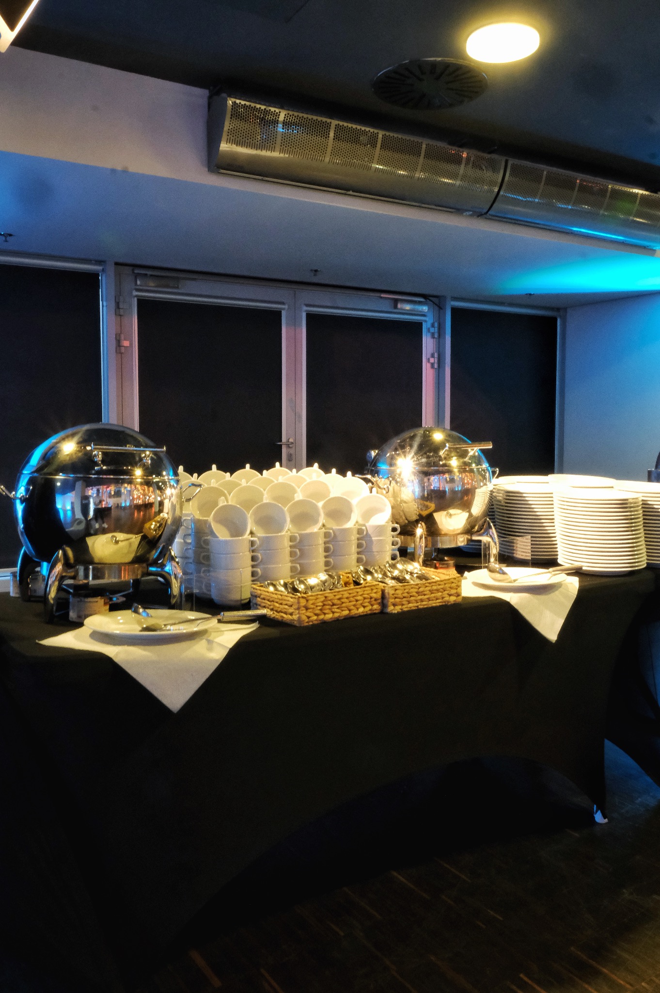 Stół z czarnym obrusem, na którym znajdują się dwa metalowe bemary, stosy białych filiżanek i talerzy, oraz koszyczki ze sztućcami, ustawione w pomieszczeniu oświetlonym niebieskim światłem.