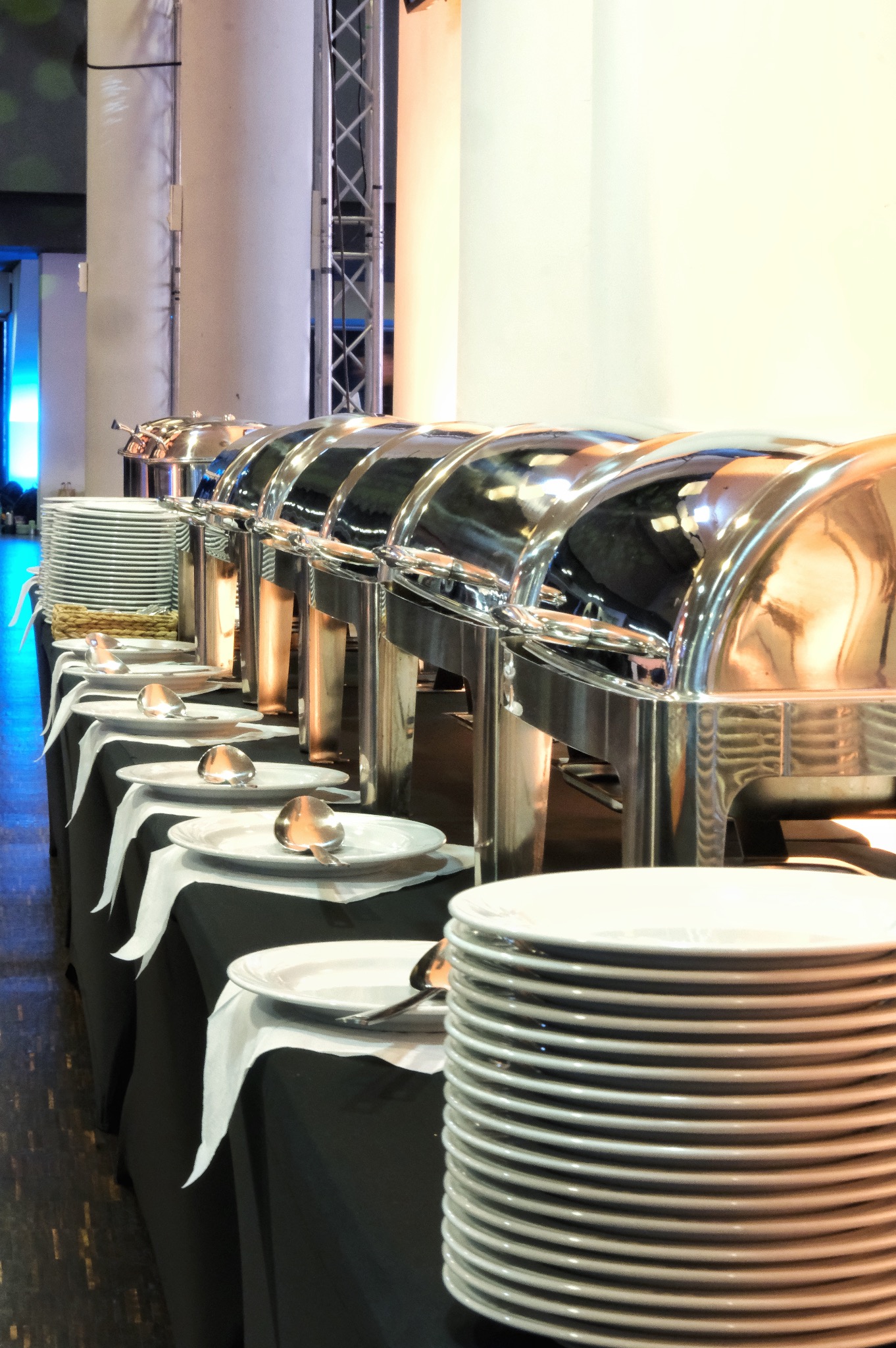 Cateringowy bufet z podgrzewaczami, ułożonymi talerzami i serwetkami na czarnym obrusie w sali eventowej, widok z perspektywy.