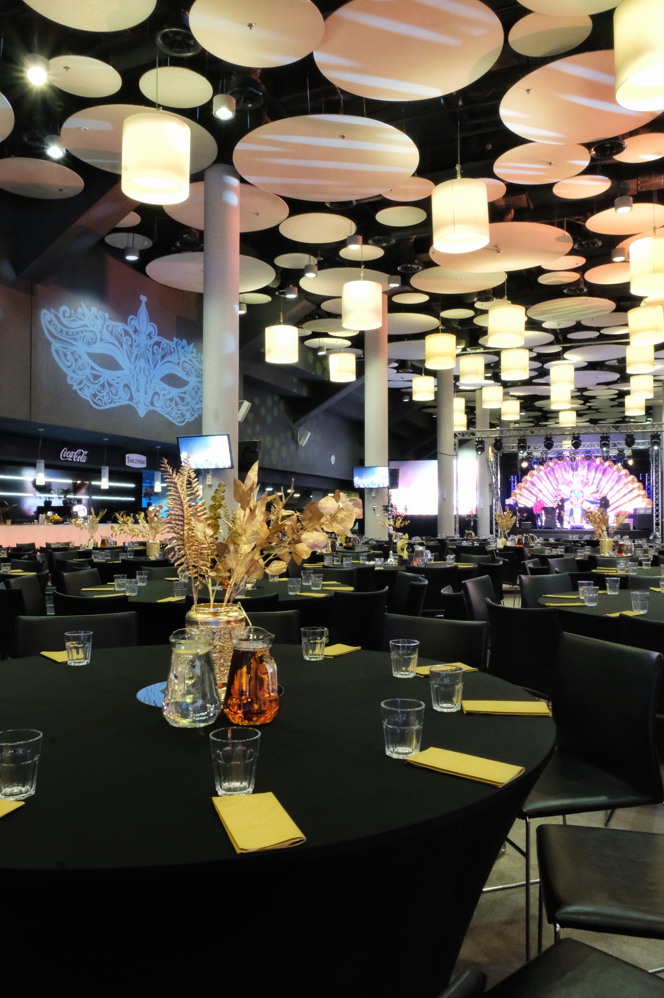 Uroczysta sala balowa z okrągłymi stołami nakrytymi czarnymi obrusami, szklankami, żółtymi serwetkami i złotymi dekoracjami kwiatowymi w wazonach. Na scenie oświetlenie i zarys sylwetek. Na ścianie...