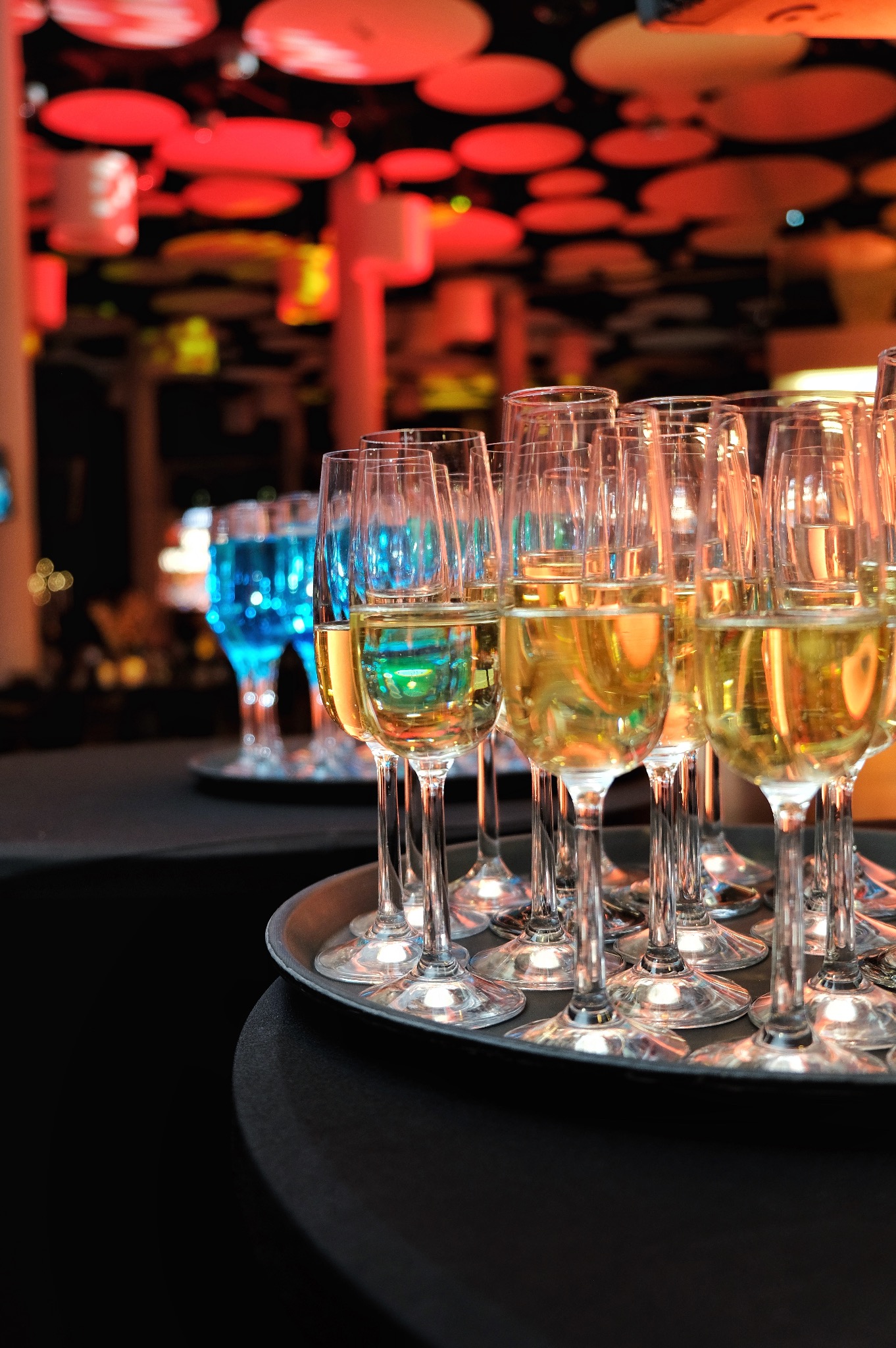 Szereg kieliszków do szampana wypełnionych złotym napojem, ustawionych na srebrnej tacy z rozmytym tłem z dekoracjami w odcieniach czerwieni i niebieskiego, sugerującymi eleganckie przyjęcie.