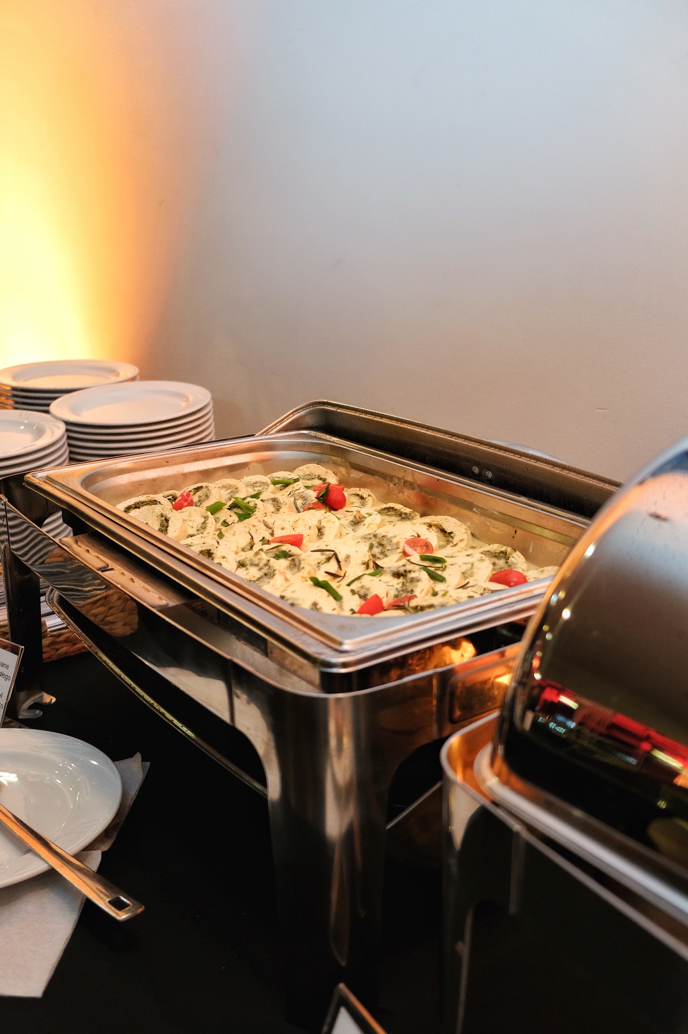 Metalowy bemar z podgrzewaną potrawą: pierożki z zielonym farszem w kremowym sosie, udekorowane kawałkami pomidorów, na stole z czarnym obrusem, obok stos białych talerzy i sztućce.