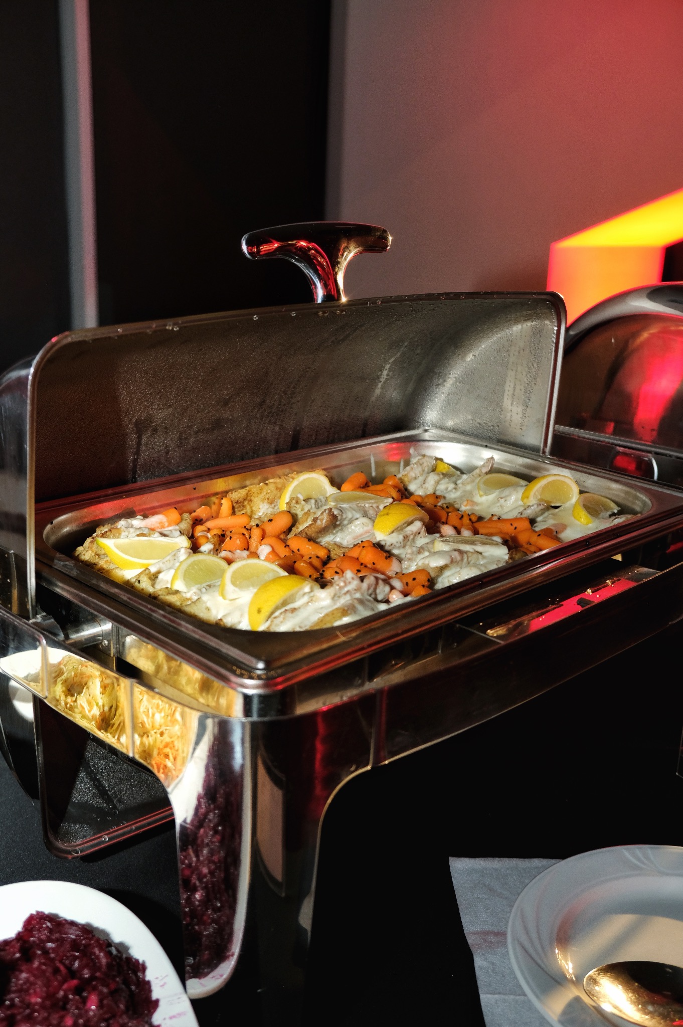 Podgrzewacz bufetowy ze srebrnej stali nierdzewnej z otwartą pokrywą, wypełniony rybą w sosie, marchewkami i plasterkami cytryny, ustawiony na czarnym stole obok talerza z czerwoną sałatką.
