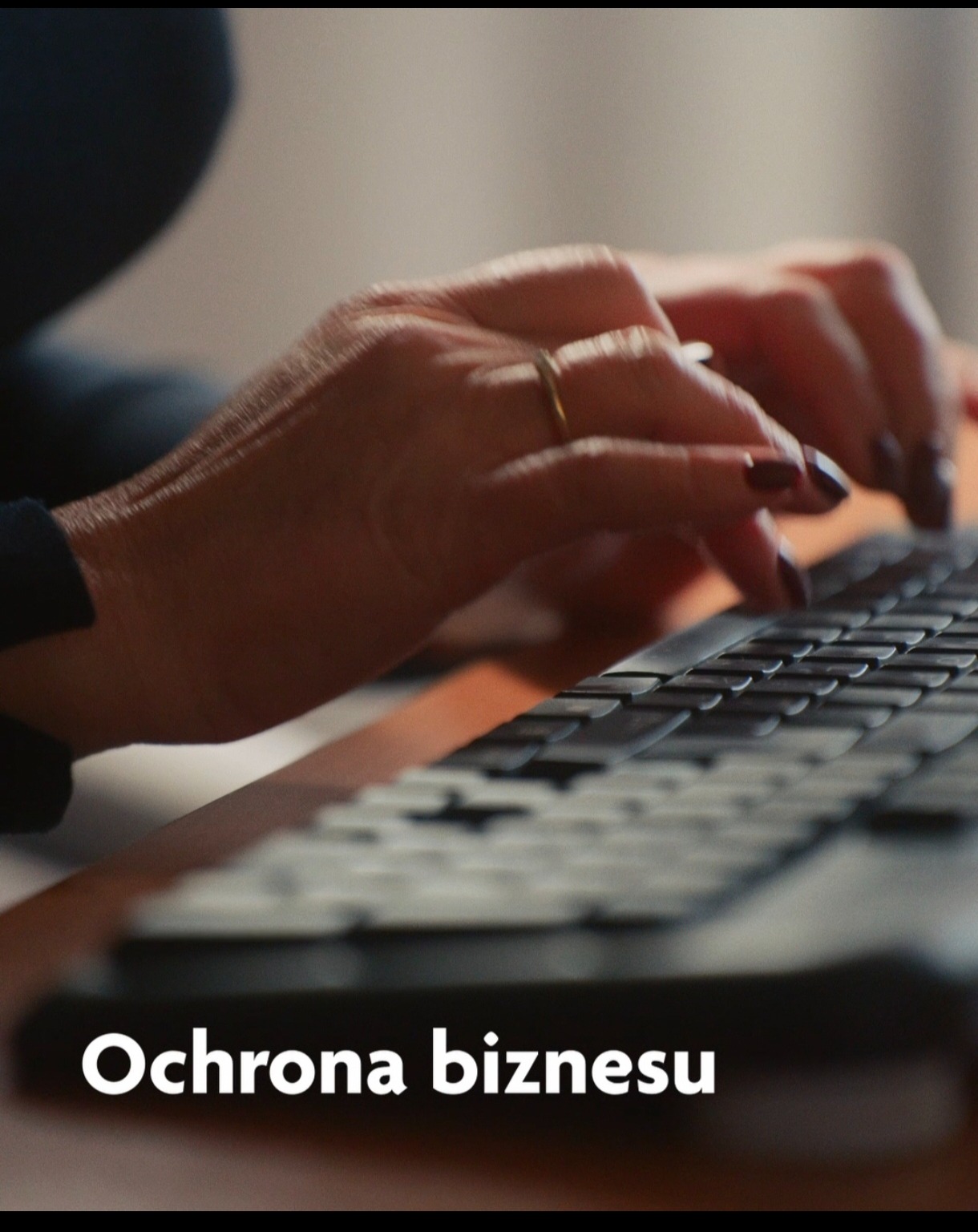 Dłonie kobiety z pierścionkiem na palcu wpisują tekst na klawiaturze komputera, oświetlonej ciepłym światłem.