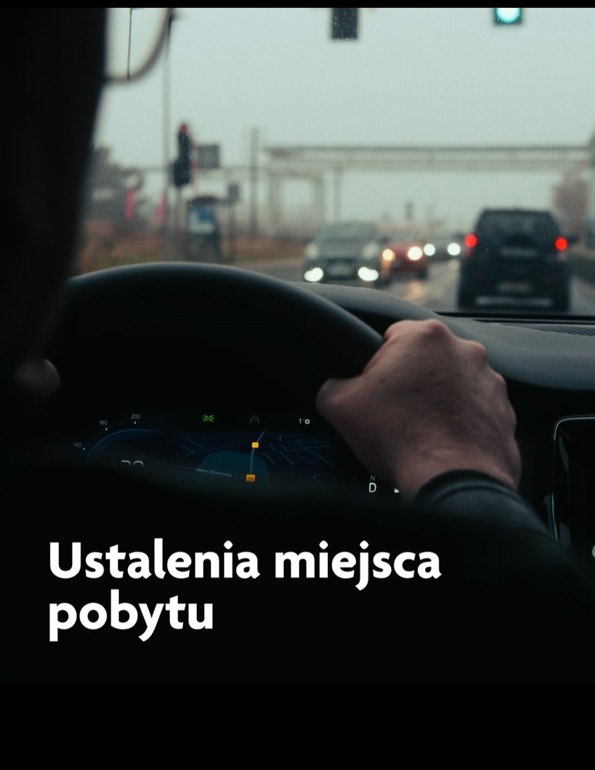 Kierowca z ręką na kierownicy w samochodzie, widok zza ramienia na drogę miejską z sygnalizacją świetlną i inne pojazdy w pochmurny dzień, na desce rozdzielczej wyświetlona mapa nawigacji.