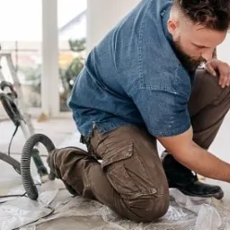 Pracownik w jeansowej koszuli i brązowych spodniach szlifuje ścianę przy podłodze, widoczna szlifierka z odkurzaczem na tle jasnego pokoju po remoncie.