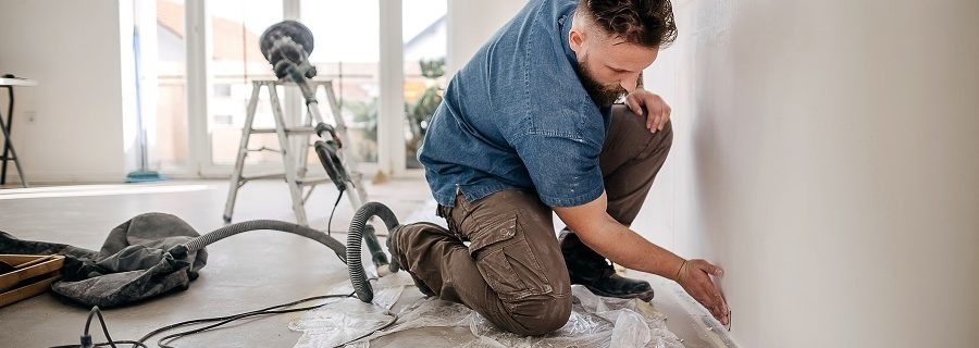 Pracownik w jeansowej koszuli i brązowych spodniach szlifuje ścianę przy podłodze, widoczna szlifierka z odkurzaczem na tle jasnego pokoju po remoncie.