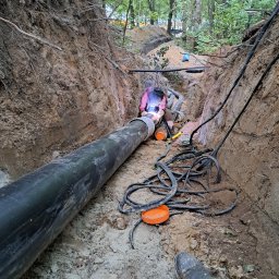 Krzysztof Barczycki Usługi Montersko-Spawalnicze - Spawanie rurociągu w wykopie ziemnym, spawacz w masce i kombinezonie, widoczne kable, rury, narzędzia i ślady spawania.