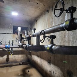 Krzysztof Barczycki Usługi Montersko-Spawalnicze - Instalacja przemysłowa z rurami, zaworami kołowymi i złączkami, umieszczona w pomieszczeniu o surowych, betonowych ścianach, częściowo zalanych wodą. Widoczne ślady korozji i zabrudzenia.