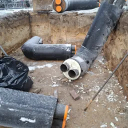 Rury ciepłownicze w wykopie, pokryte szronem, z widoczną izolacją i elementami montażowymi, przygotowane do spawania.