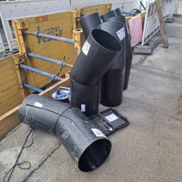 Krzysztof Barczycki Usługi Montersko-Spawalnicze - Czarne rury kanalizacyjne z widocznymi etykietami i śladami po cięciu, ułożone obok wykopu zabezpieczonego metalowymi panelami i rozpórkami.