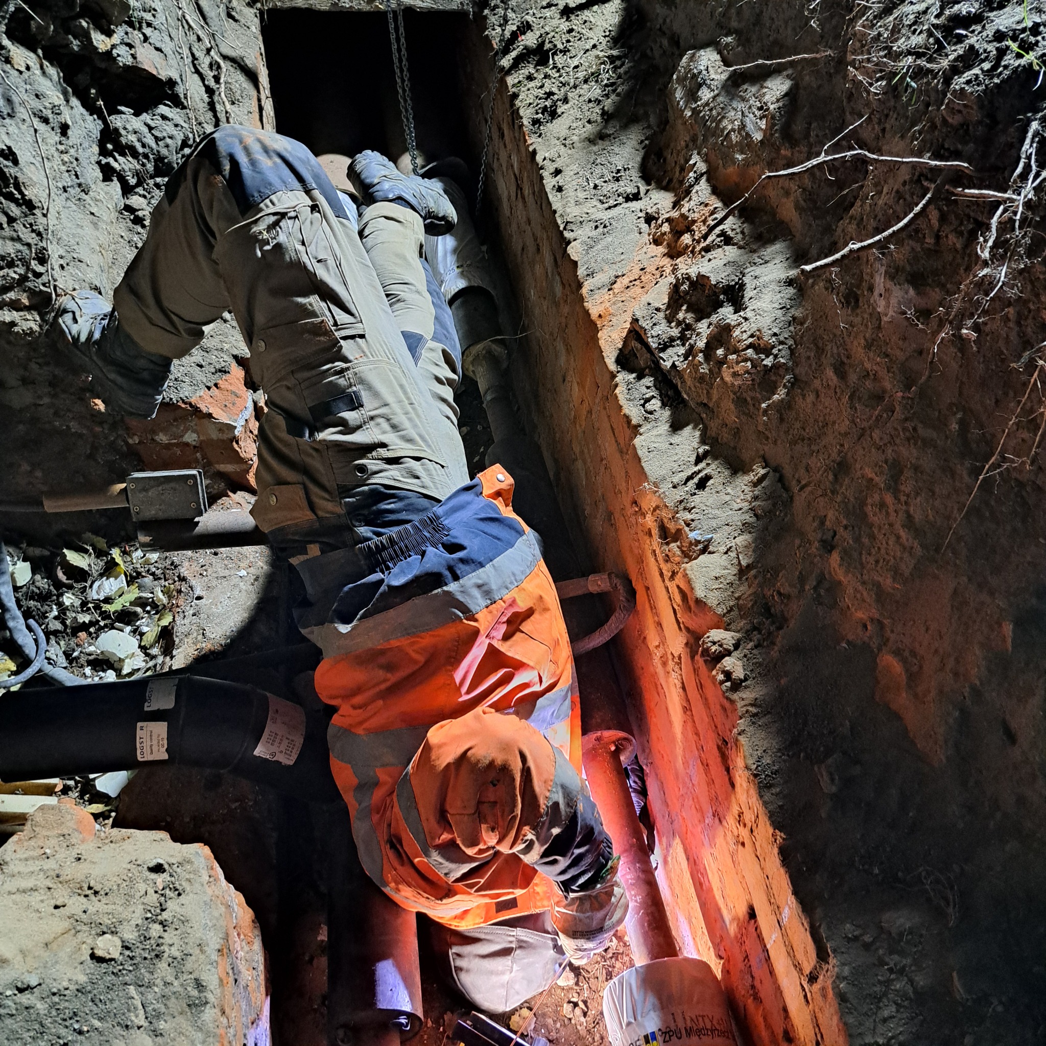 Pracownik w pomarańczowej kamizelce w wykopie, prawdopodobnie podczas prac spawalniczych na rurach, widoczne iskry i ciemne otoczenie wykopu.