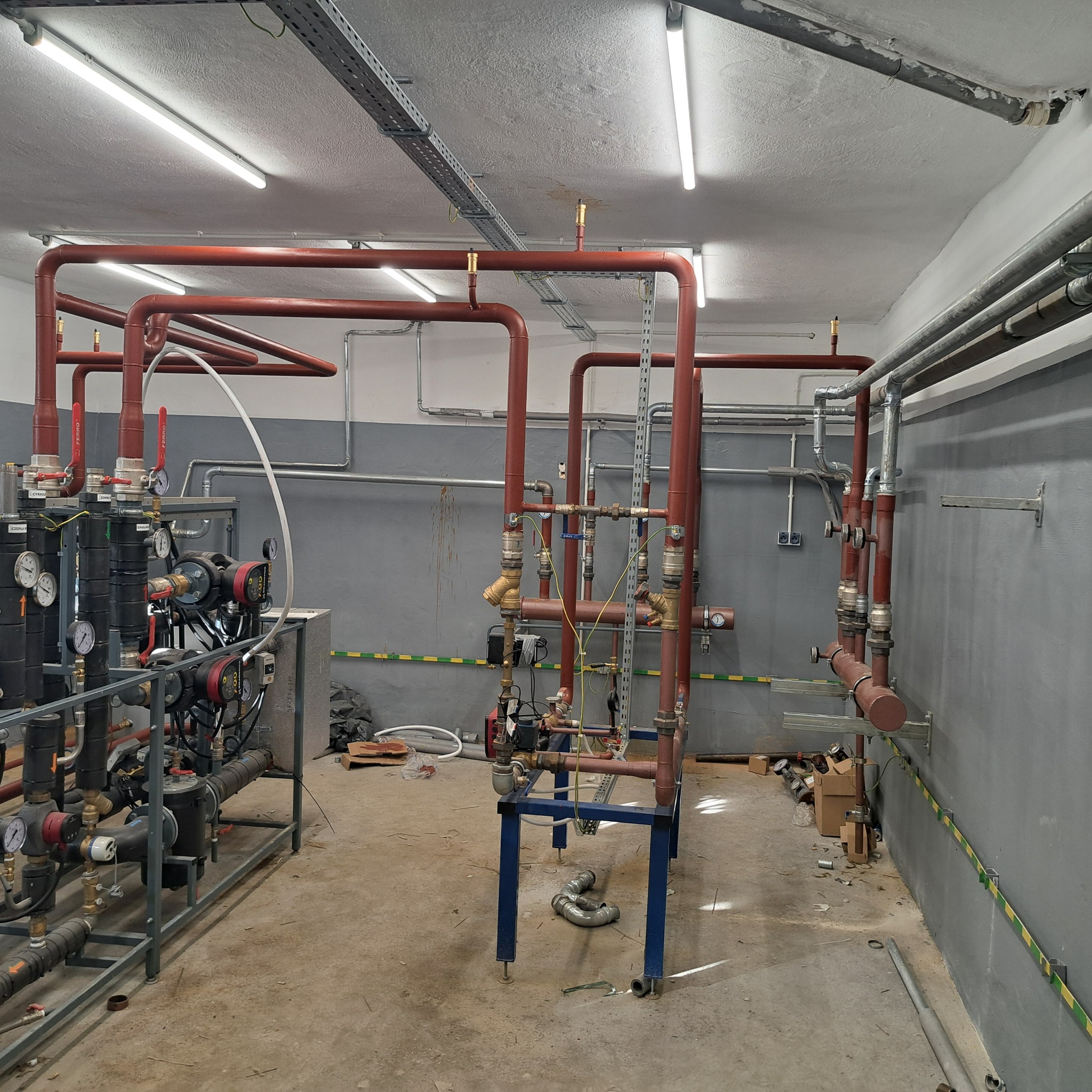 Rozbudowana instalacja centralnego ogrzewania z rurami w kolorze ceglastej czerwieni, zaworami i manometrami, zamontowana w pomieszczeniu technicznym z szarymi ścianami i betonową podłogą.