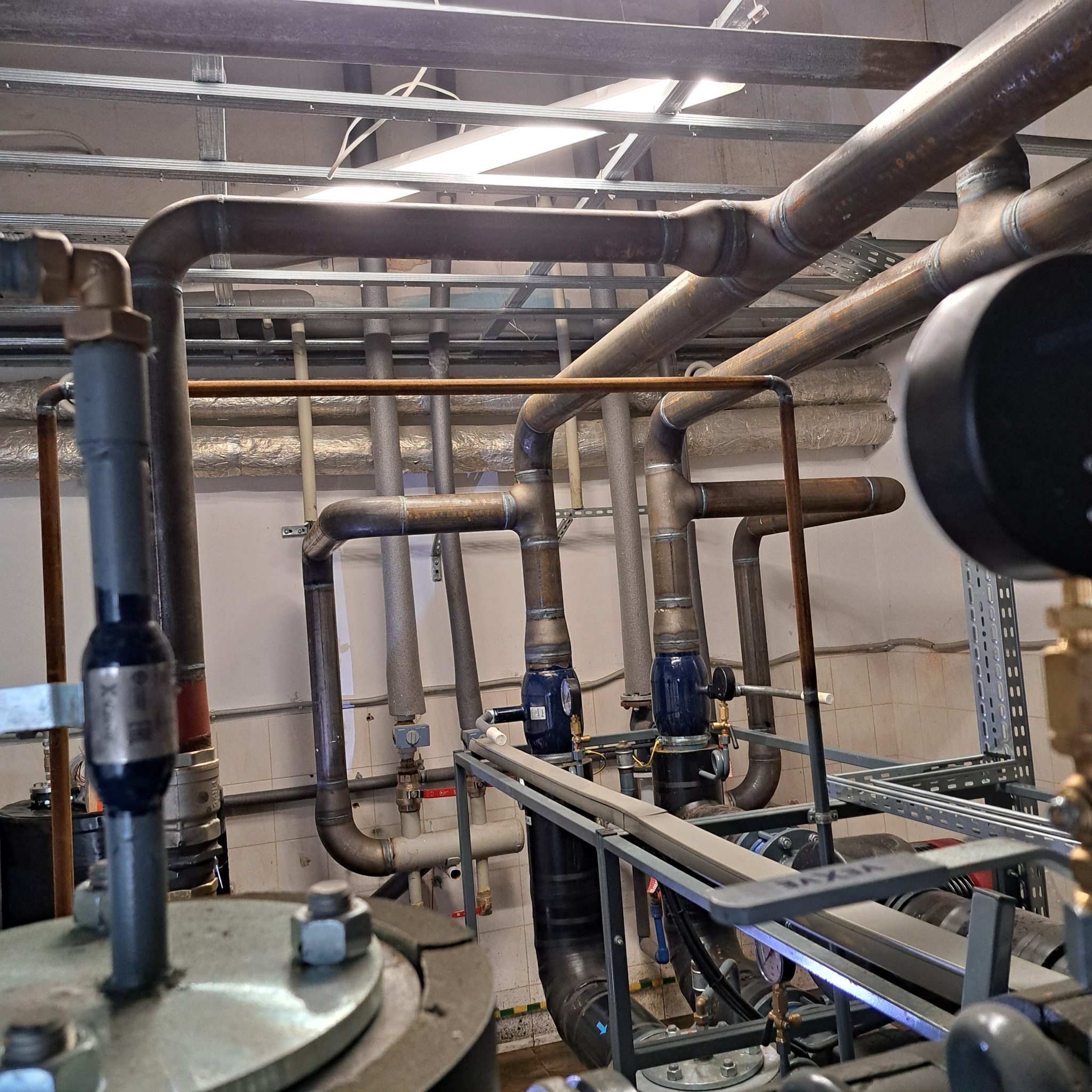Kompleksowy system rur miedzianych i stalowych z zaworami i manometrami, zainstalowany pod sufitem w pomieszczeniu technicznym, z widocznymi spawami na łączeniach rur.
