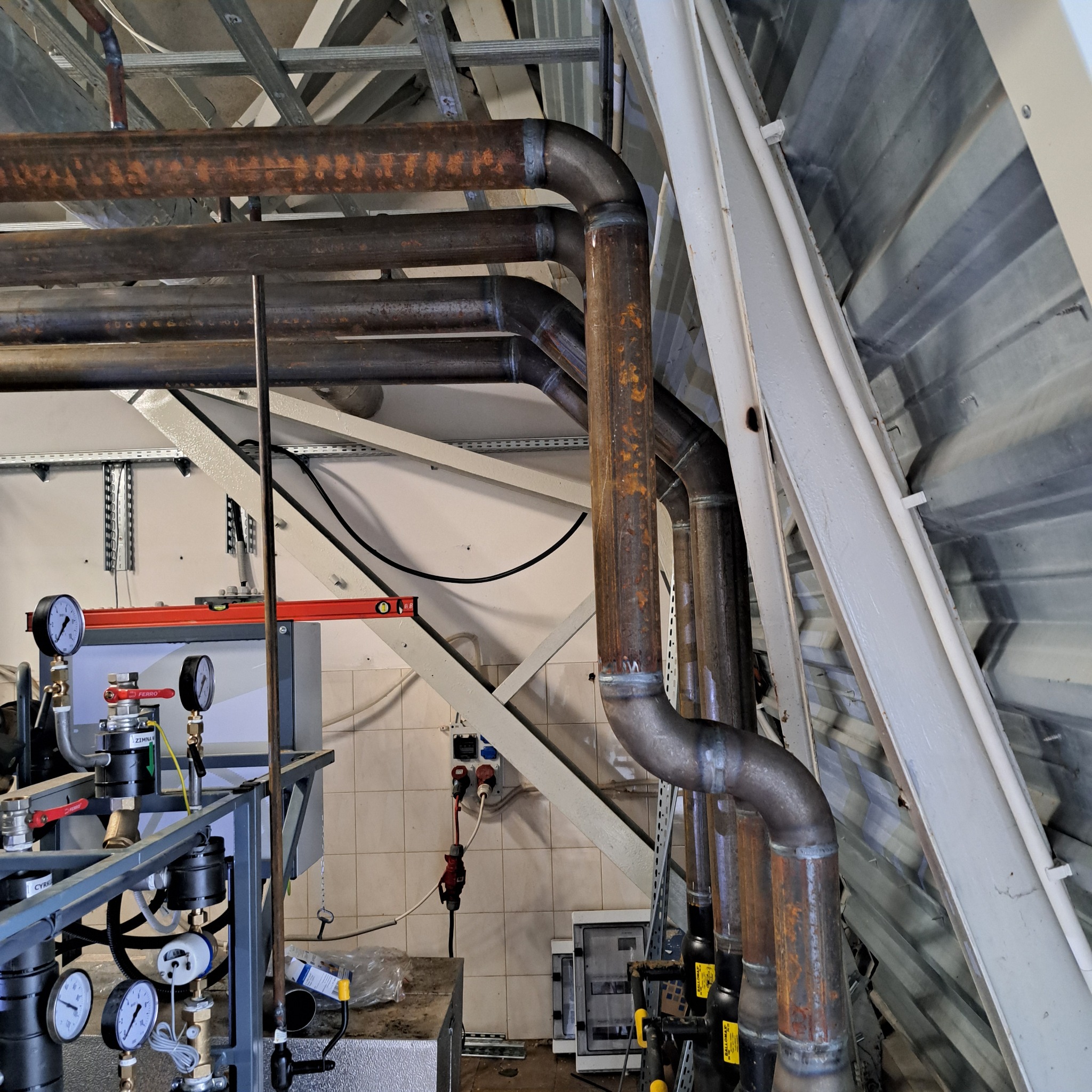 Rdzewiejące metalowe rury z widocznymi spawami, część instalacji przemysłowej z manometrami i zaworami, na tle konstrukcji stalowej i białych płytek