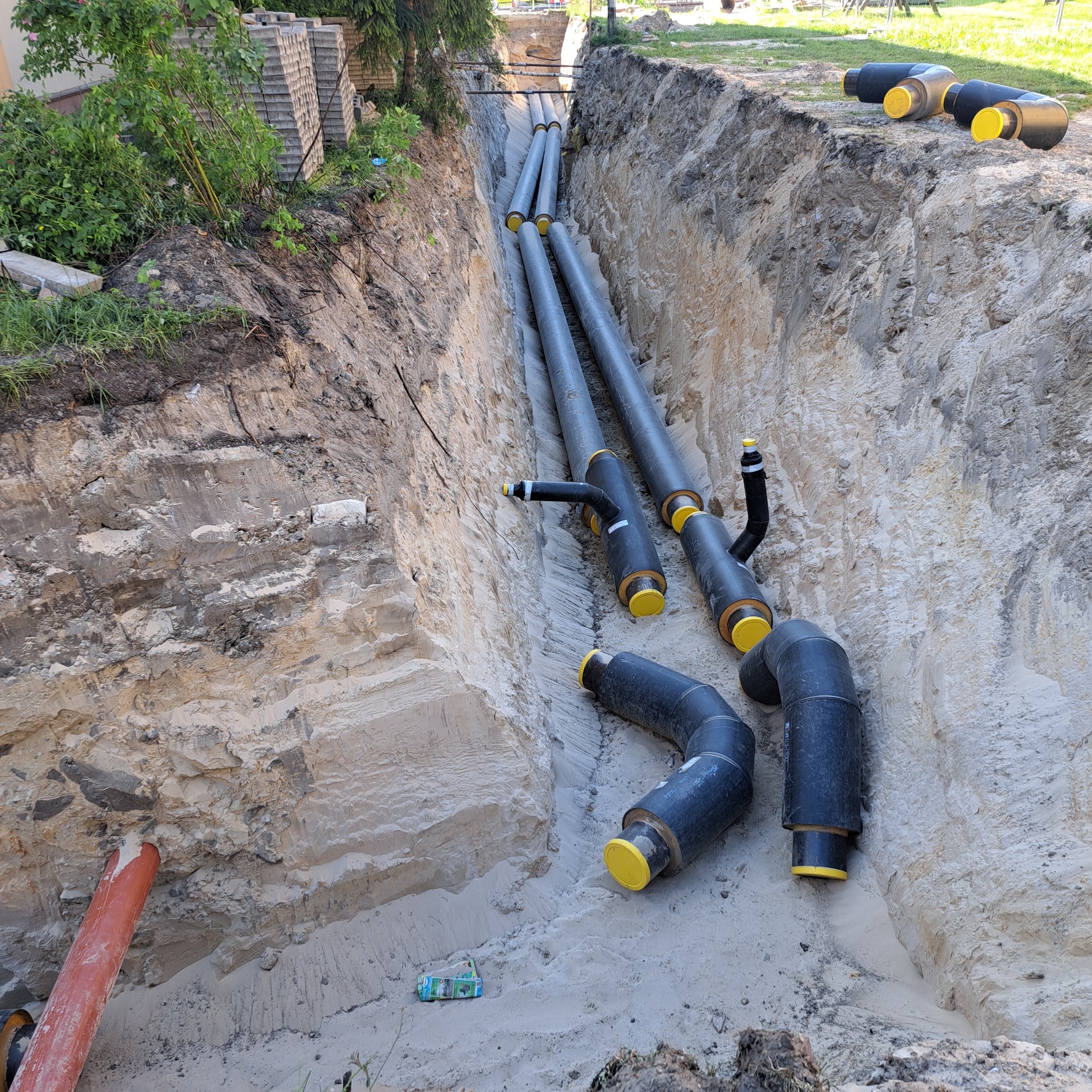 Ułożone w wykopie rury ciepłownicze z żółtymi zaślepkami, widoczny przekrój ziemi z warstwami piasku i gliny, w tle trawnik i budynek.