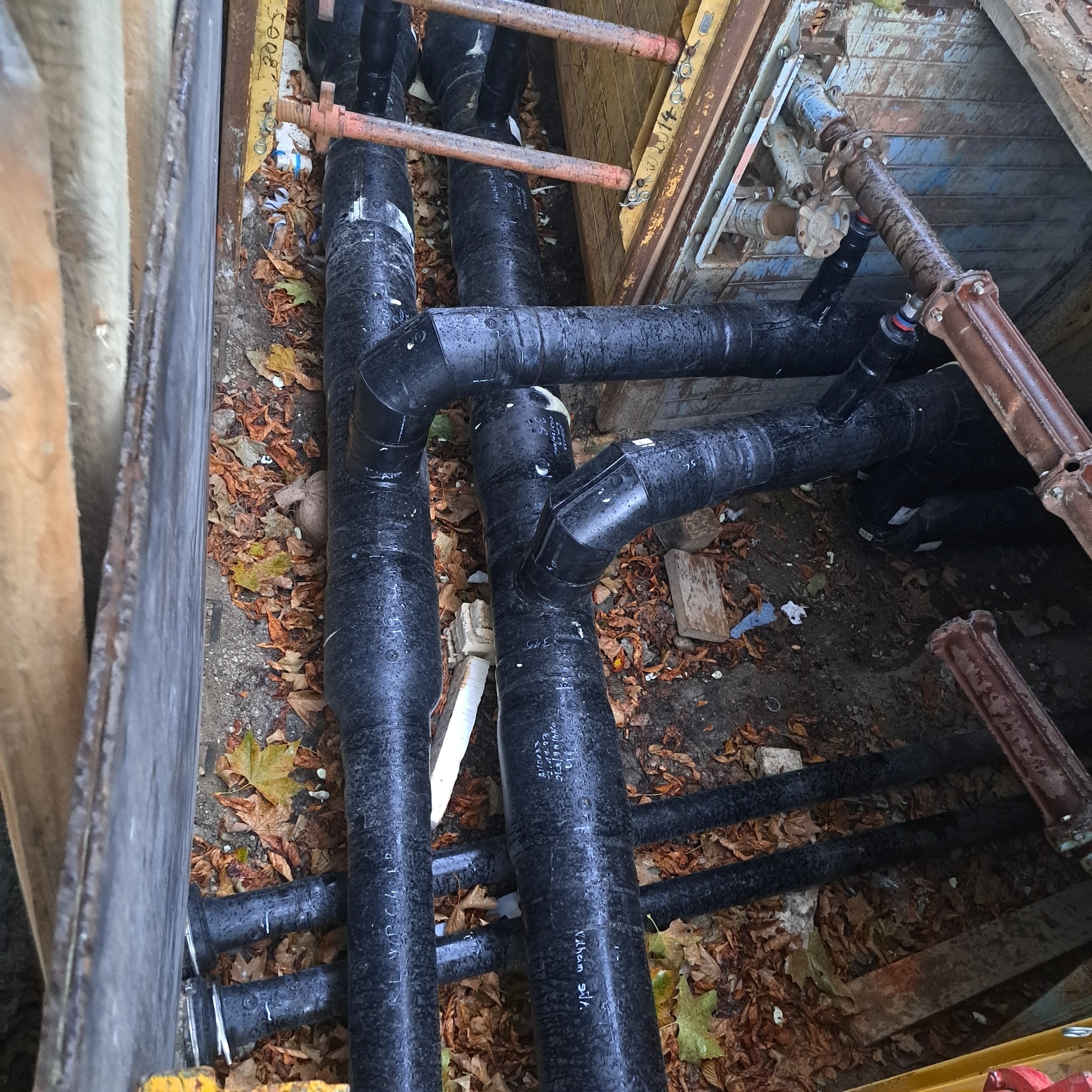 Złożony system czarnych rur kanalizacyjnych ułożonych w wykopie zabezpieczonym metalowymi szalunkami, widoczne liście na dnie wykopu.