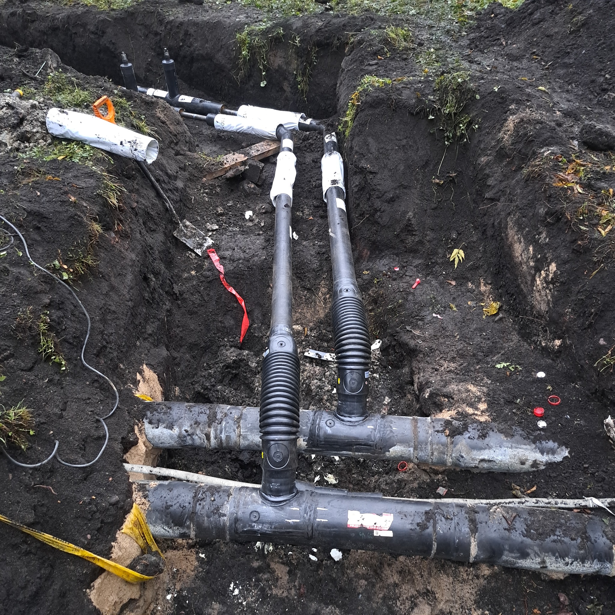 Instalacja podziemna: czarne rury z karbowanymi złączkami w wykopie ziemnym, częściowo owinięte białą folią, obok narzędzia i taśma ostrzegawcza.