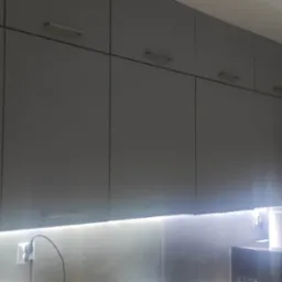 Szereg szarych szafek kuchennych wiszących z oświetleniem LED podszafkowym, widoczny fragment ściany z gniazdkiem elektrycznym i przewodem, z boku lodówka.