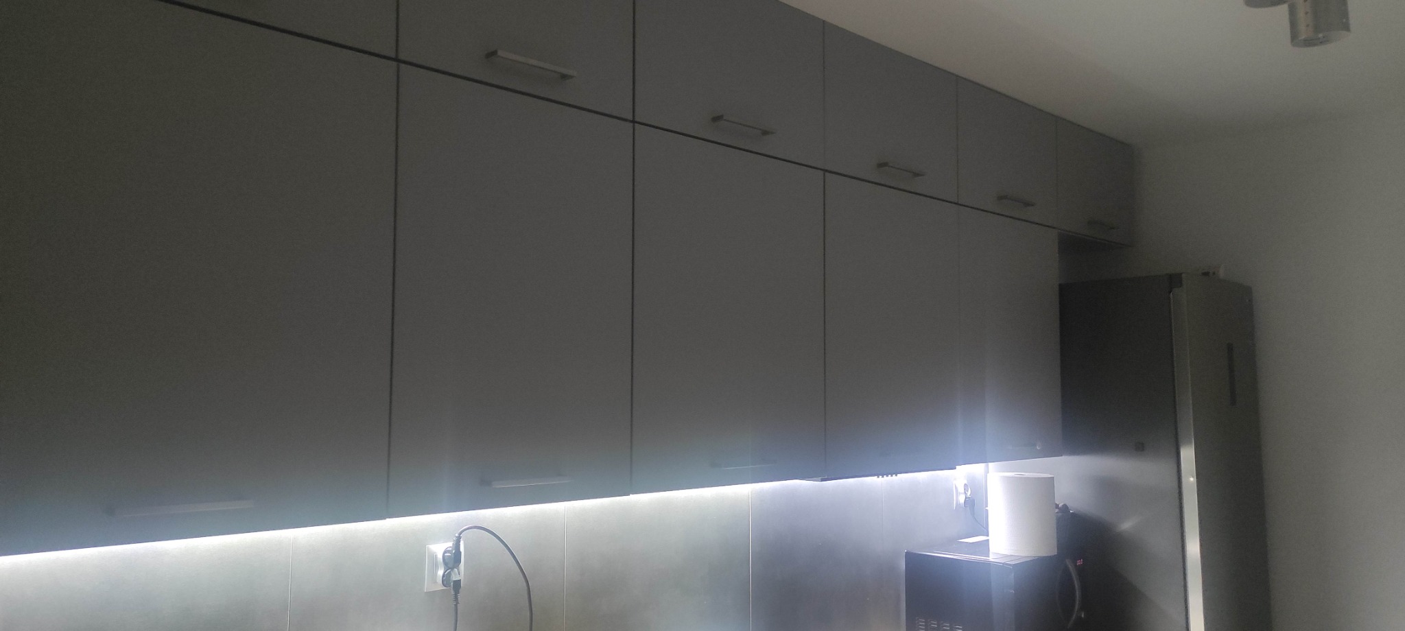 Szereg szarych szafek kuchennych wiszących z oświetleniem LED podszafkowym, widoczny fragment ściany z gniazdkiem elektrycznym i przewodem, z boku lodówka.