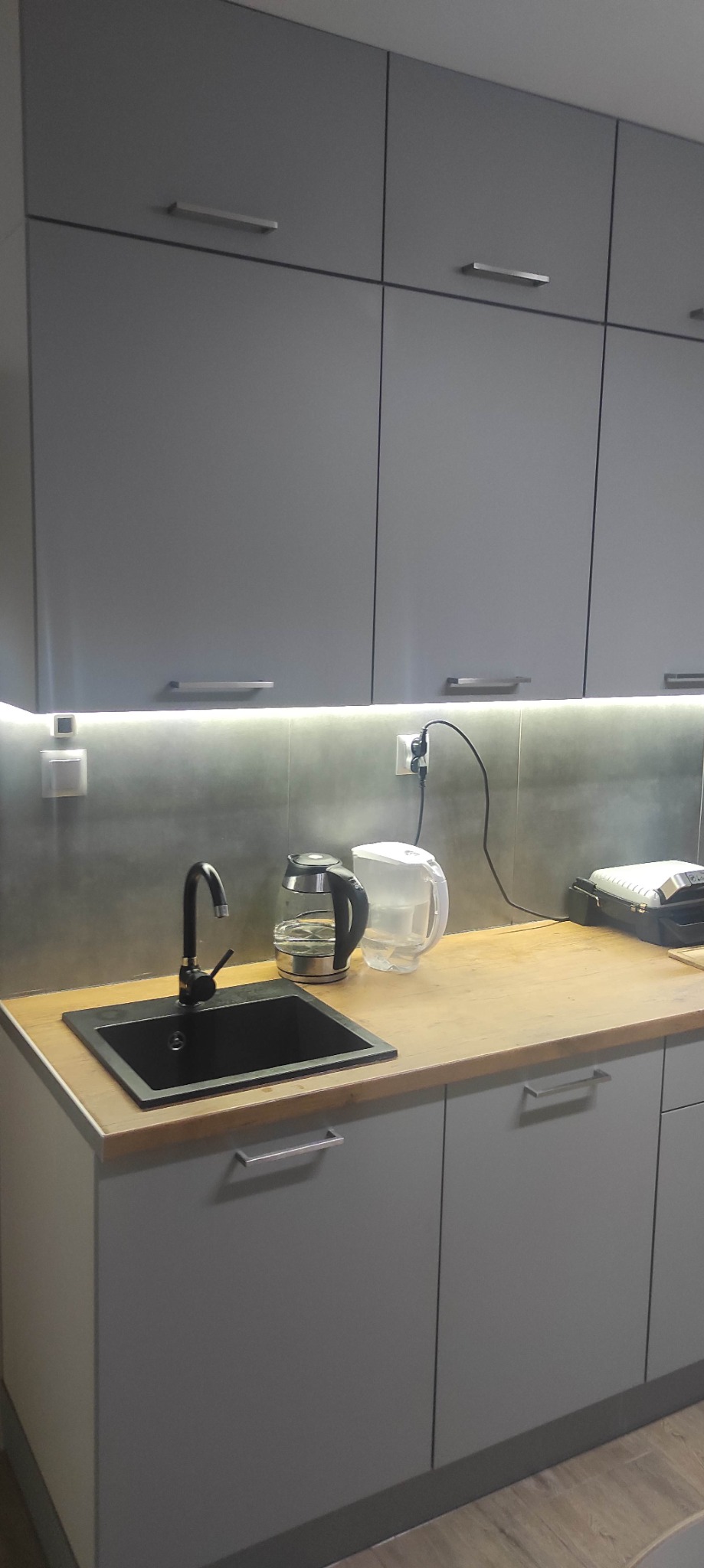 Szare szafki kuchenne z drewnianym blatem, czarnym zlewem i baterią, podświetlone listwą LED, z widocznymi gniazdkami elektrycznymi i czajnikiem elektrycznym.