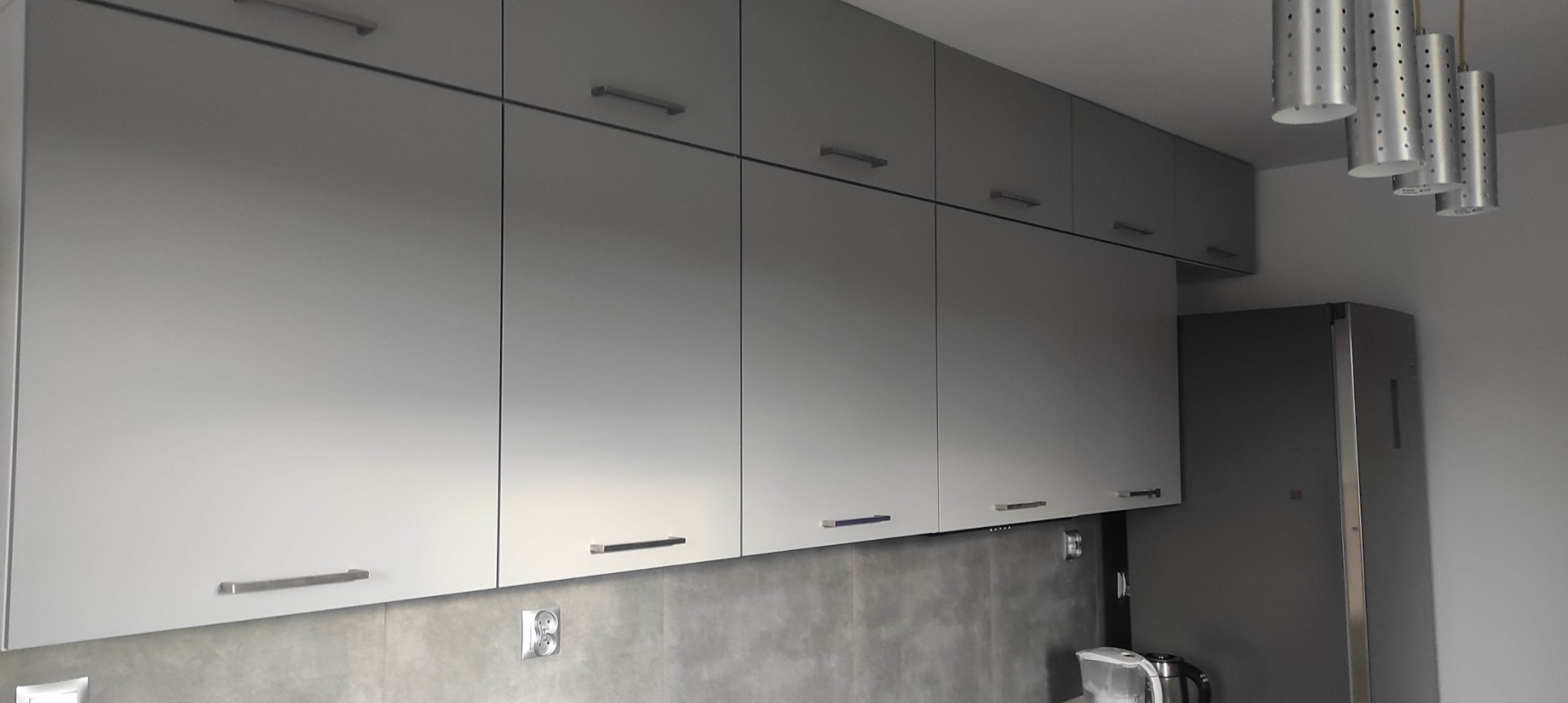 Szare szafki kuchenne wiszące z podłużnymi uchwytami, betonowa ściana nad blatem, widoczne gniazdko elektryczne i fragment lodówki oraz designerska lampa sufitowa.