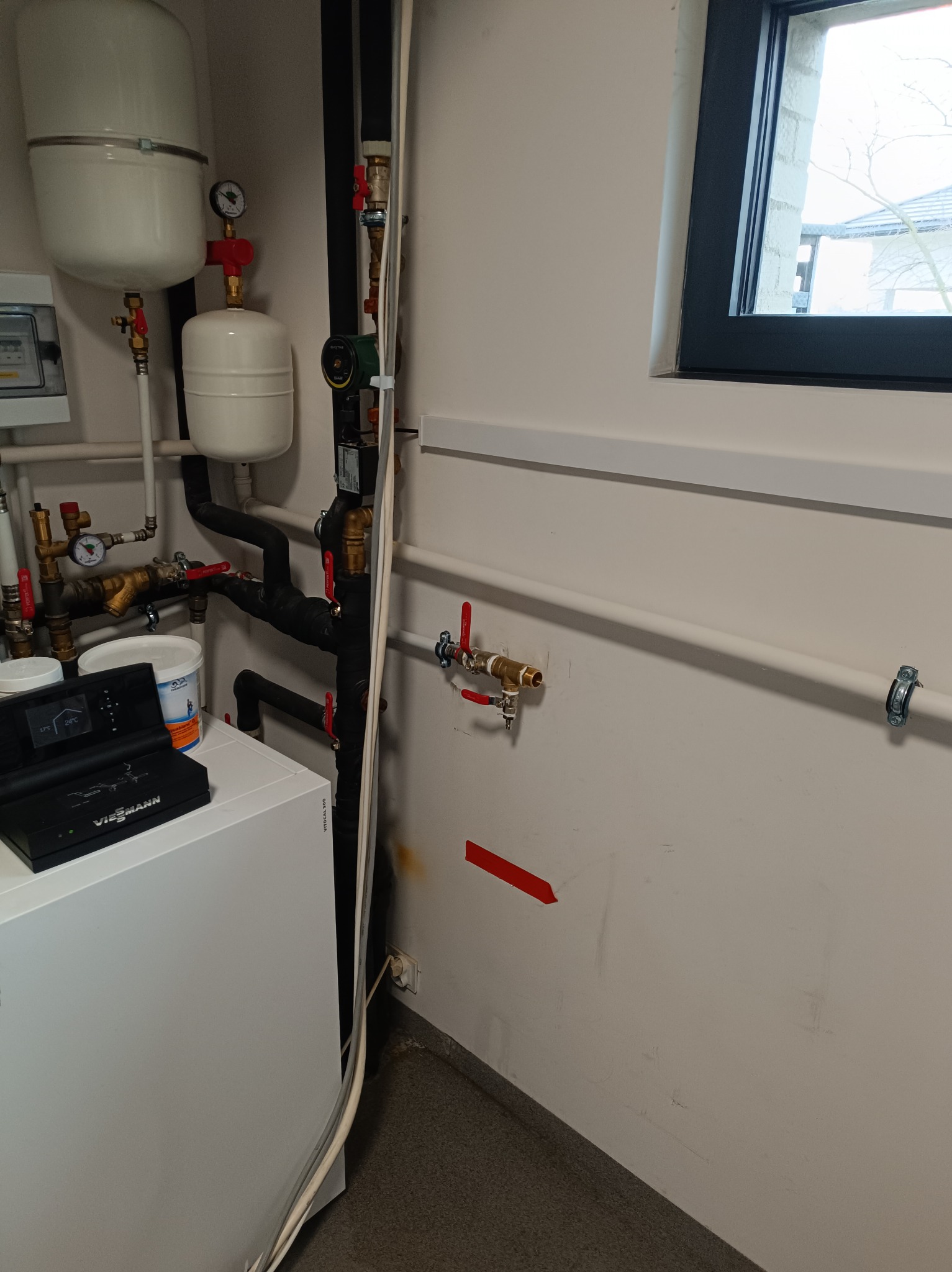 Biały piec Viessmann z czarnym panelem sterowania, zbiorniki wyrównawcze, rury instalacji CO i okno w tle. Widoczne zawory i manometry.