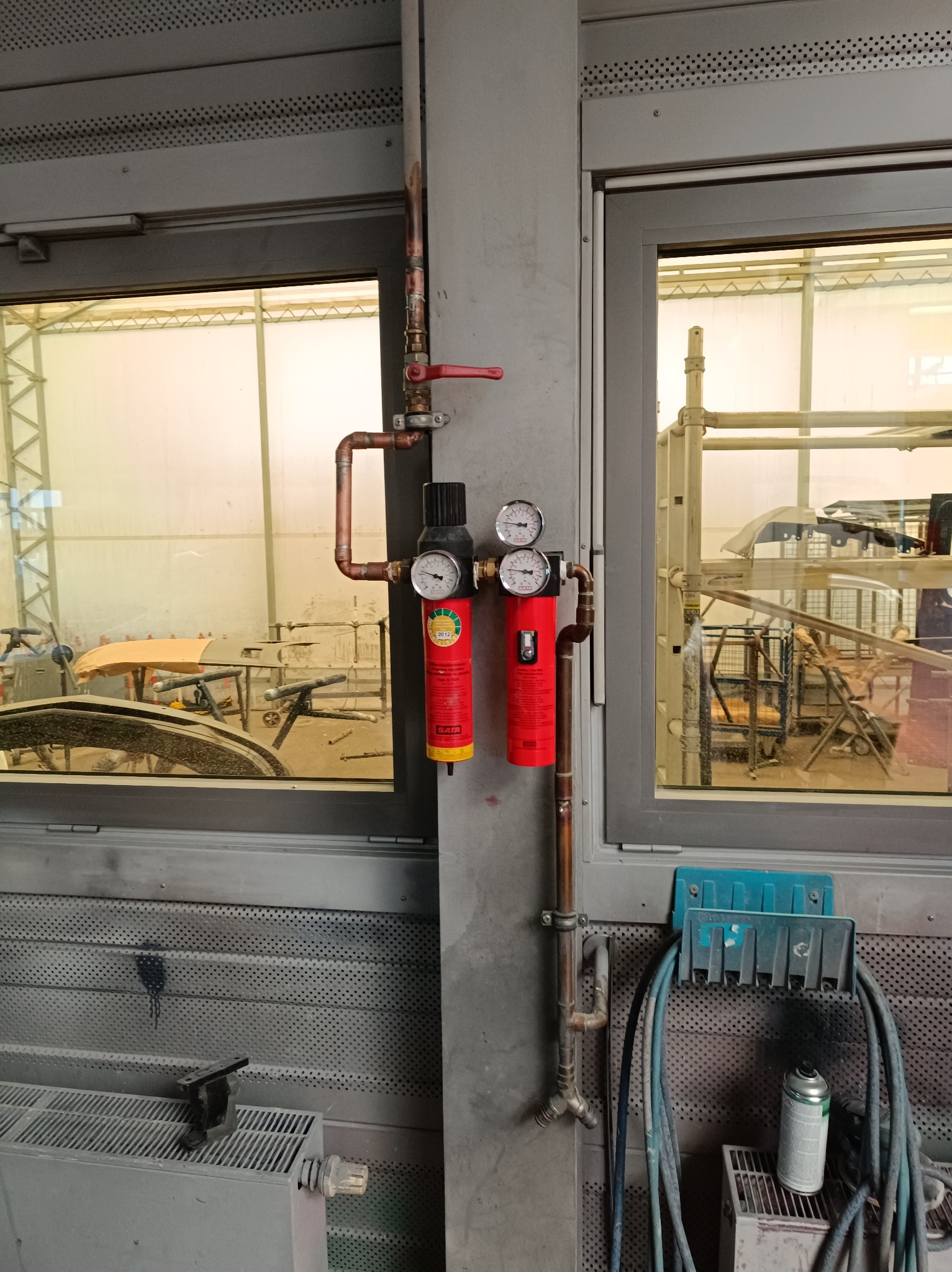 Instalacja pneumatyczna z filtrami i manometrami w lakierni samochodowej, widoczne miedziane rury i akcesoria, w tle elementy karoserii samochodowej.