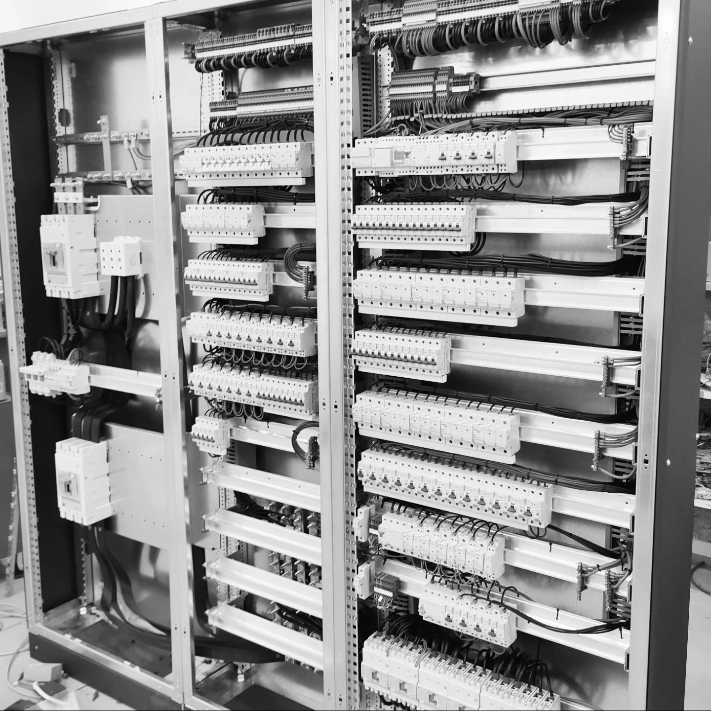 Rozdzielnia elektryczna w szafie sterowniczej, widoczne liczne bezpieczniki i okablowanie wewnątrz metalowej obudowy. Czarno-biała tonacja podkreśla techniczną naturę instalacji.