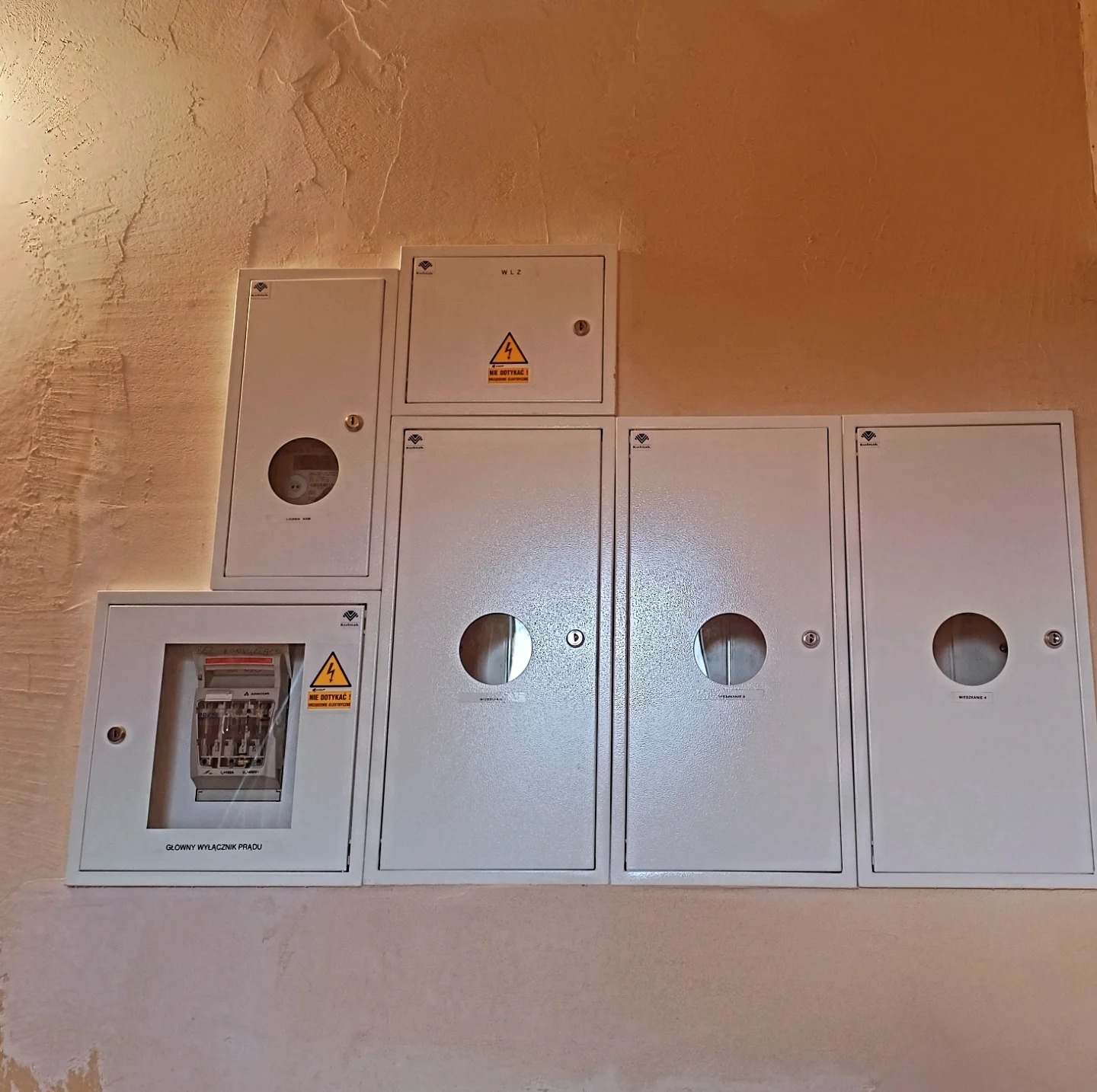 Zestaw białych skrzynek elektrycznych (liczniki, wyłączniki) na beżowej ścianie o fakturze baranka. Jedna skrzynka z widocznym licznikiem i napisem 'Główny wyłącznik prądu'.