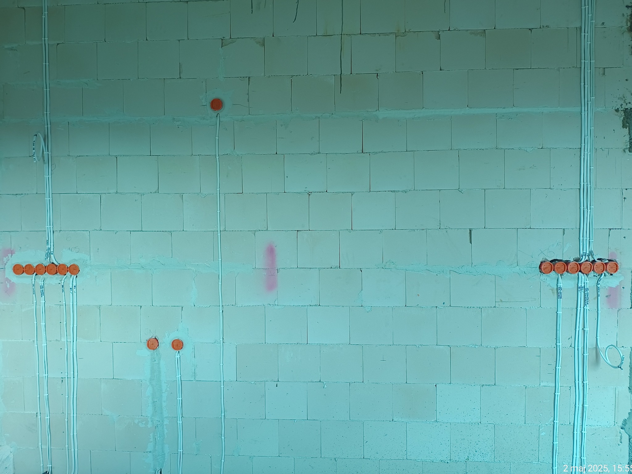 Instalacja elektryczna w trakcie montażu: białe przewody prowadzone po ścianie z bloczków, pomarańczowe puszki elektryczne. Widoczne napisy na przewodach.