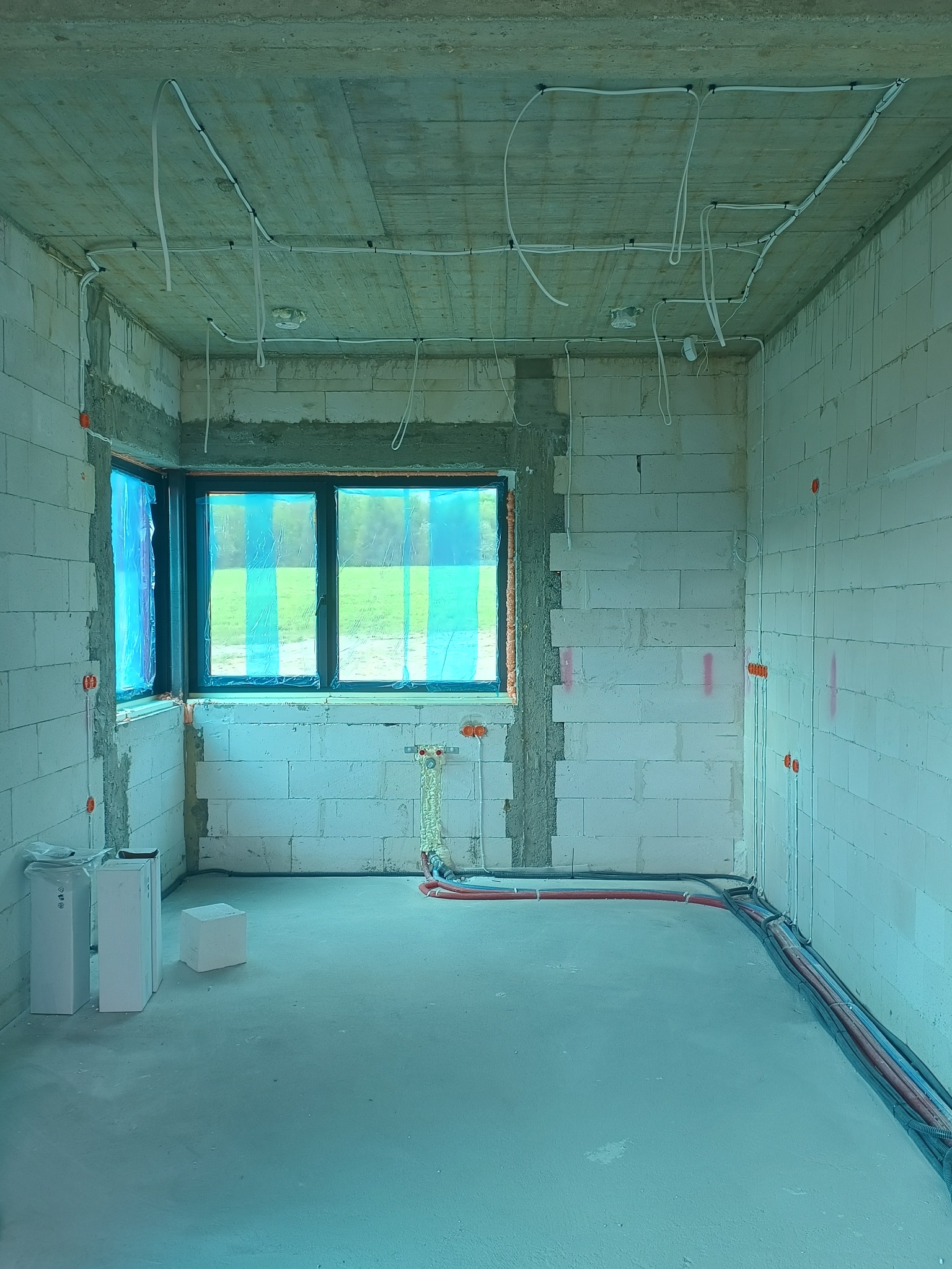Surowy stan wnętrza z instalacją elektryczną i rurami grzewczymi na ścianach i suficie, okno zabezpieczone folią, bloczki betonowe, surowy betonowy strop.