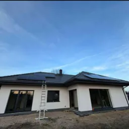 Montaż paneli słonecznych na dachu domu jednorodzinnego z ciemną dachówką. Aluminiowa drabina oparta o dach. Błękitne niebo w tle.