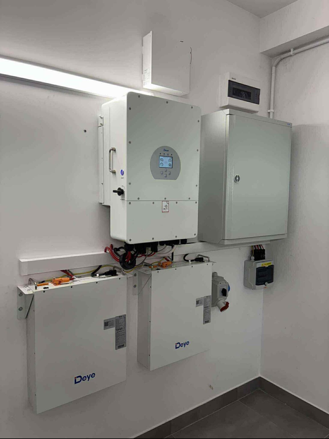 Białe akumulatory i falownik Deye zamontowane na ścianie w pomieszczeniu. Widoczne przewody, skrzynka elektryczna i listwa oświetleniowa.