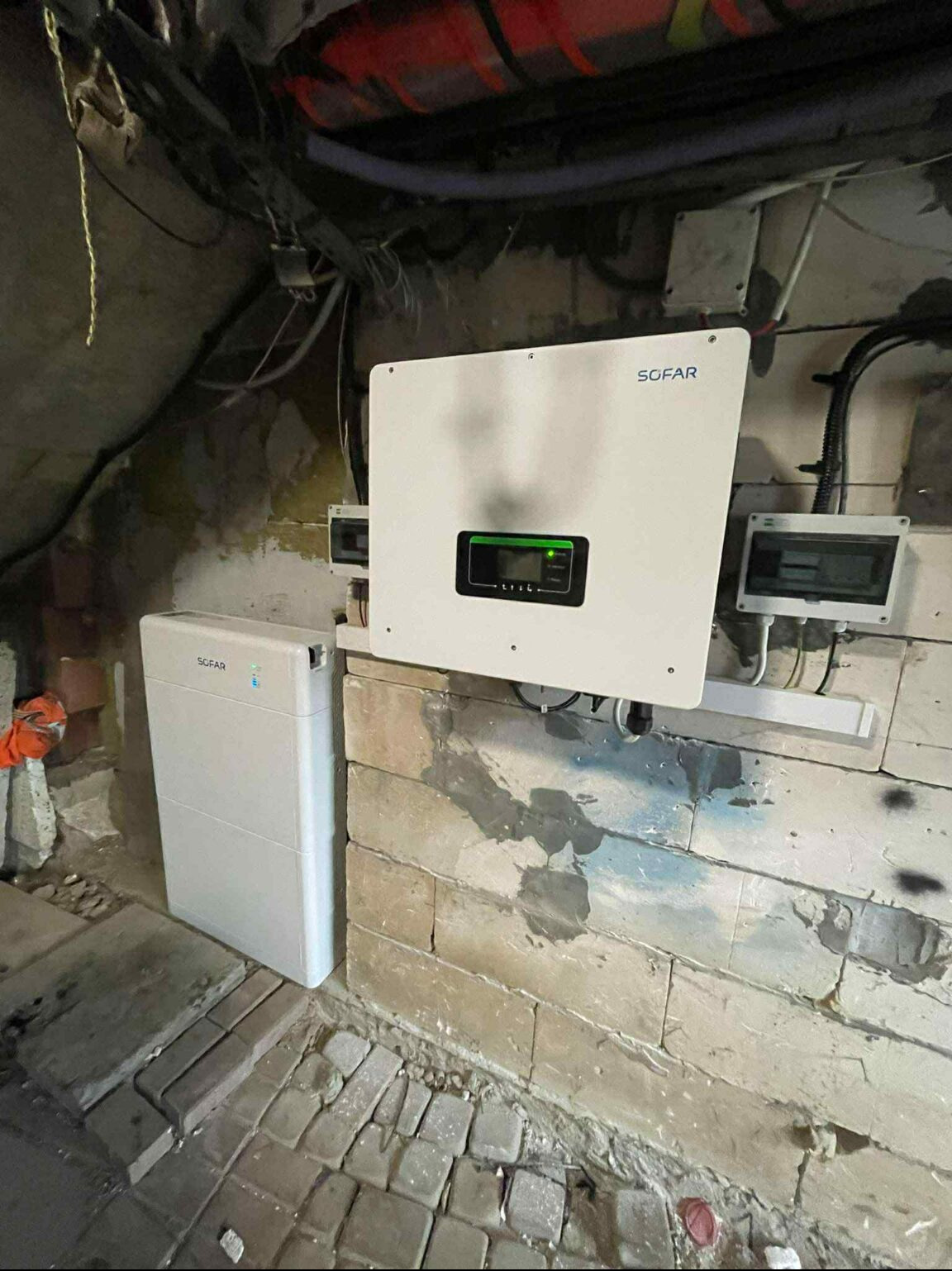 Biały inwerter Sofar i magazyn energii Sofar zamontowane na ceglanej ścianie w piwnicy. Widoczne kable i instalacje elektryczne. Podłoże z kostki brukowej.