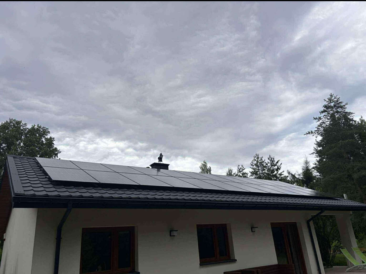 Czarny dach z panelami słonecznymi na białym domu, widok z dołu w pochmurny dzień. Panele ułożone równomiernie, komin z figurką na szczycie.