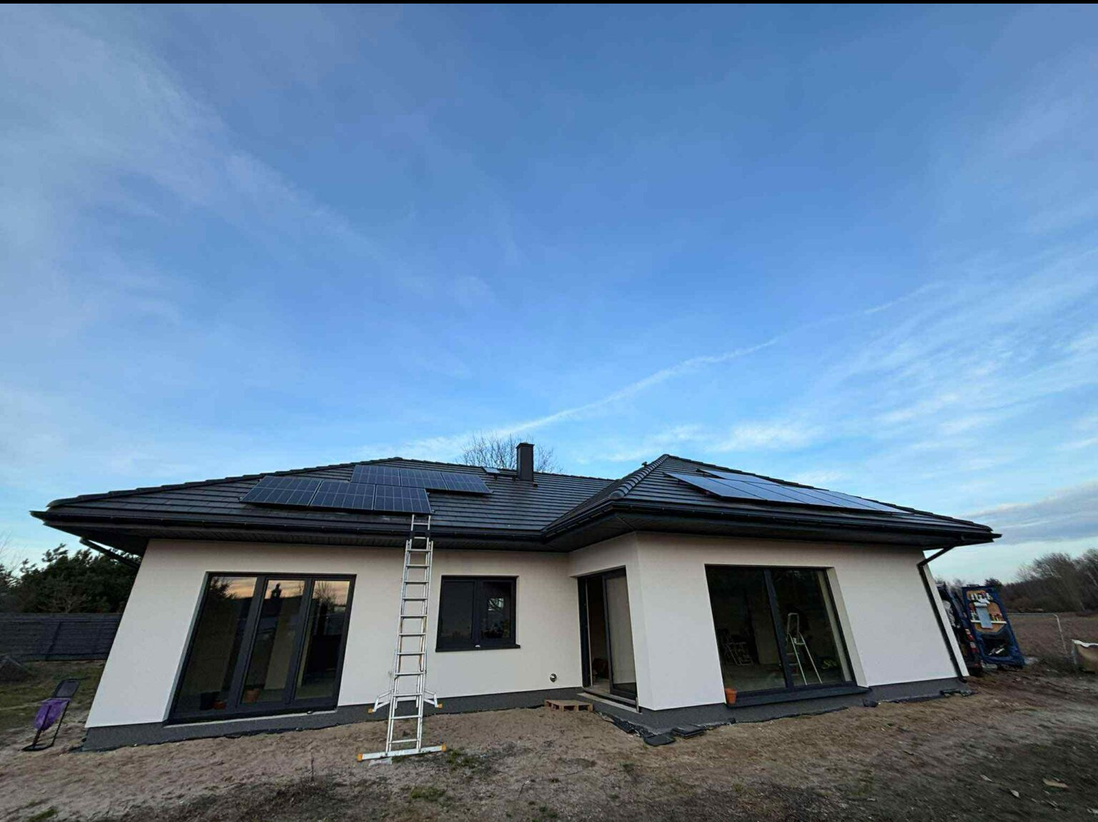 Montaż paneli słonecznych na dachu domu jednorodzinnego z ciemną dachówką. Aluminiowa drabina oparta o dach. Błękitne niebo w tle.