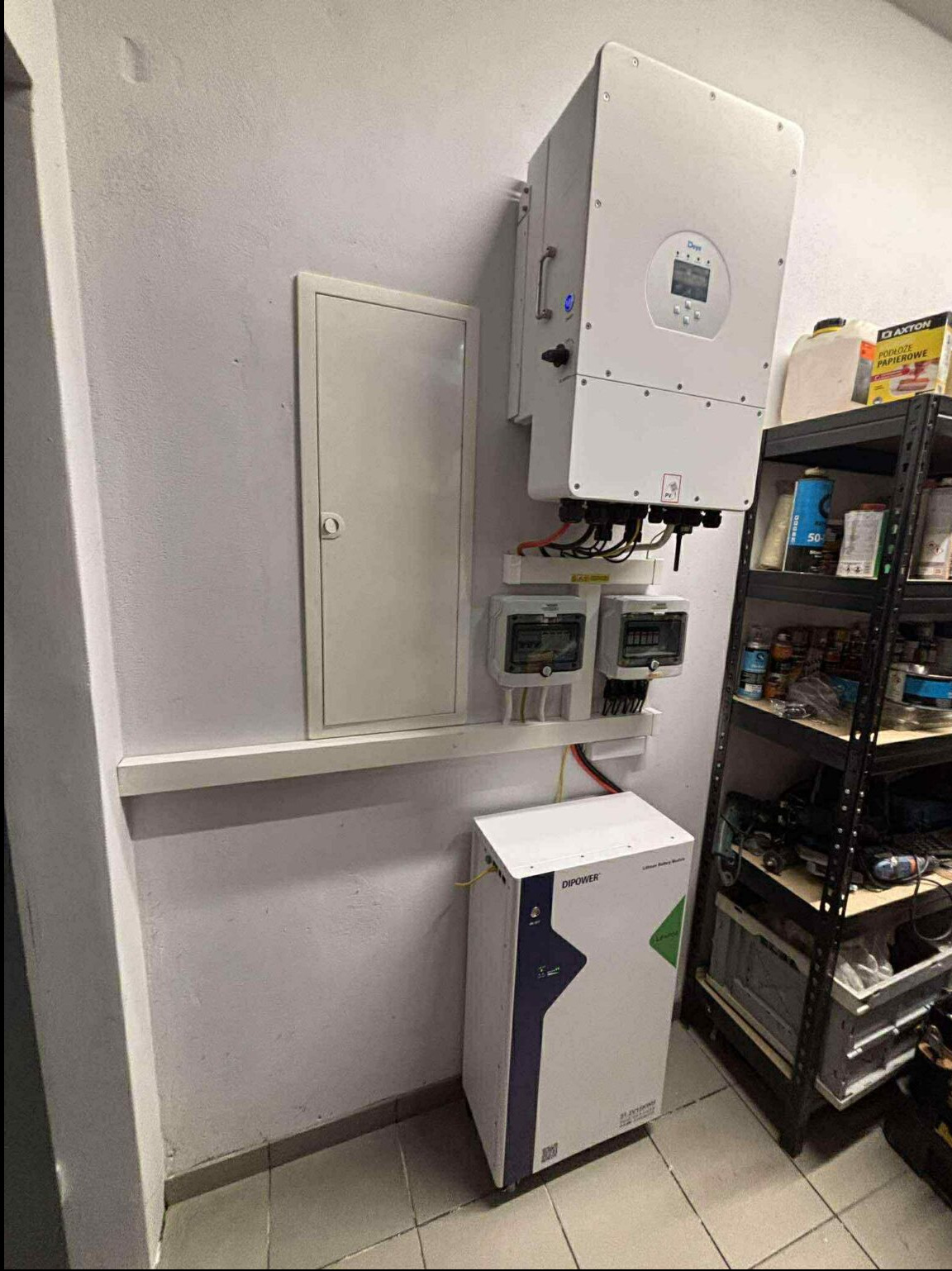 Kompletna instalacja: inwerter na ścianie, liczniki, skrzynka, obok biały akumulator DIPOWER. Półka z narzędziami i akcesoriami w tle.