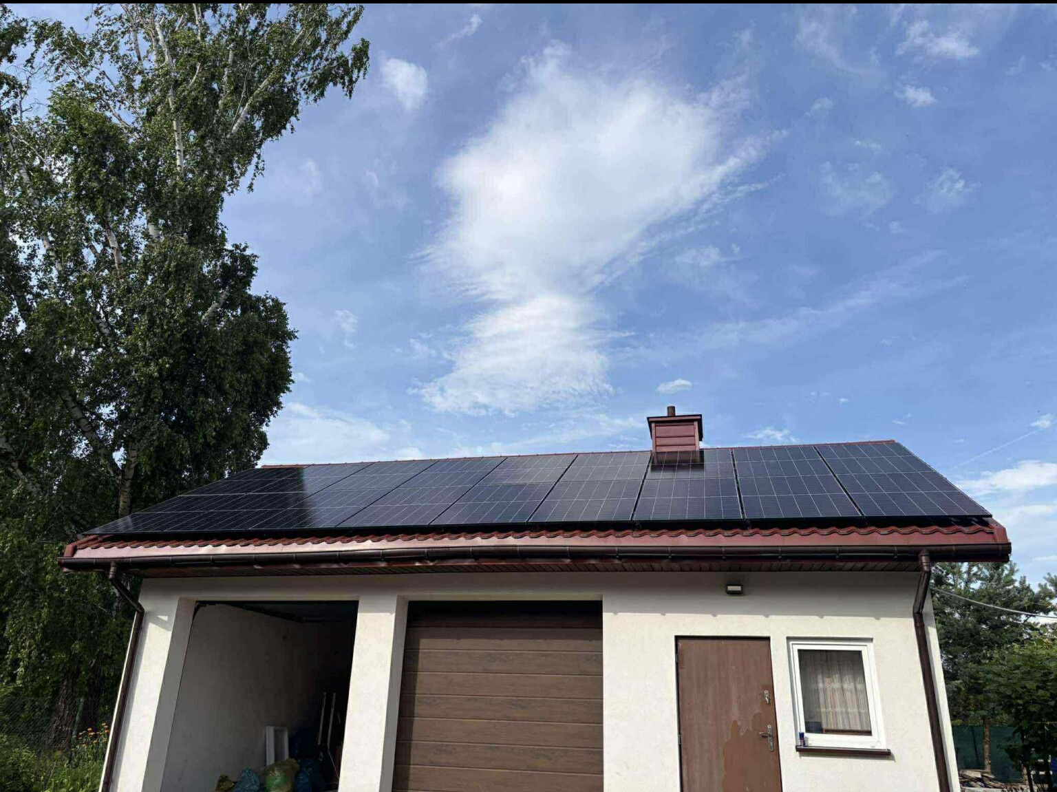Panele słoneczne na dachu garażu z brązową blachodachówką, błękitne niebo z chmurami w tle. Widok z dołu, drzewo po lewej stronie.