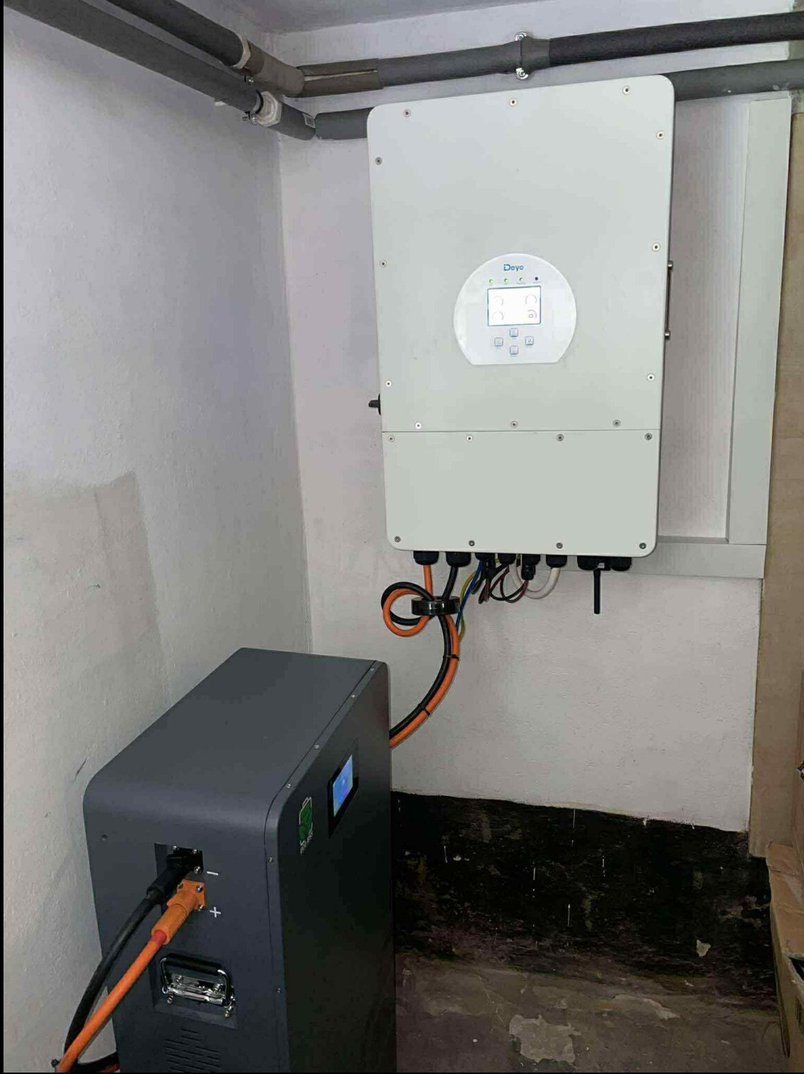 Szary magazyn energii podłączony pomarańczowymi kablami do białego inwertera na ścianie w piwnicy. Widoczne rury i czarna podłoga.