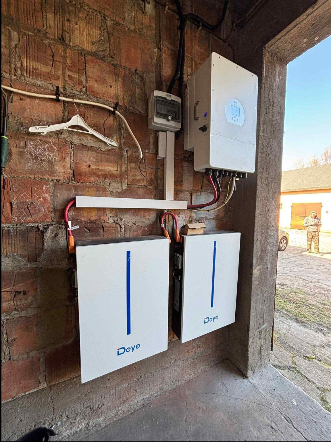 Dwa białe akumulatory Deye z niebieskimi paskami, zamontowane na ceglanej ścianie obok białego inwertera i rozdzielnicy. Widoczny fragment budynku i osoba w tle.