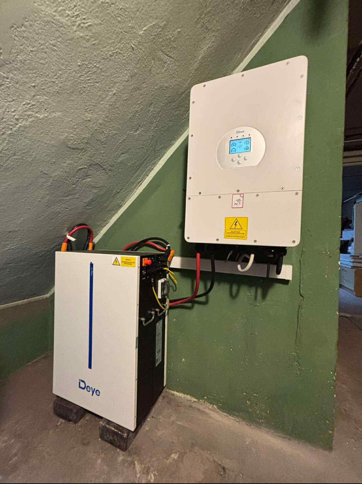 Instalacja magazynu energii Deye w Jastrzębiu-Zdroju. Widoczny akumulator na cegłach i falownik na ścianie, połączone kablami. Ściany w odcieniach zieleni.