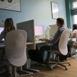 Jasne biuro z trzema osobami pracującymi przy biurkach. Monitory z danymi, nowoczesne krzesła i naturalne światło z okna tworzą profesjonalne środowisko pracy.