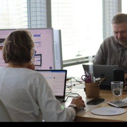 Dwie osoby pracują w biurze przy komputerach. Na biurku kalkulator, szklanka wody i przybory biurowe. W tle okno z żaluzjami i logo firmy na monitorze.