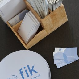 FIK FINANSE I KSIĘGOWOŚĆ SPÓŁKA Z OGRANICZONĄ ODPOWIEDZIALNOŚCIĄ - Biurko z drewnianym organizerem wypełnionym notatnikami, ołówkami i wizytówkami firmy Fik Finanse i Księgowość. Na blacie leżą podkładki z logo firmy.