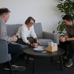 Spotkanie biznesowe w jasnym wnętrzu: trzech konsultantów omawia dokumenty przy okrągłym stole. Minimalistyczny wystrój, roślina doniczkowa w tle.