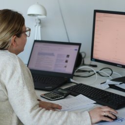 FIK FINANSE I KSIĘGOWOŚĆ SPÓŁKA Z OGRANICZONĄ ODPOWIEDZIALNOŚCIĄ - Kobieta w biurze, z laptopem, monitorem, kalkulatorem i dokumentami na biurku. Praca biurowa, analiza danych, obliczenia. Jasne, naturalne oświetlenie.