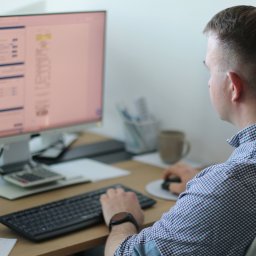 FIK FINANSE I KSIĘGOWOŚĆ SPÓŁKA Z OGRANICZONĄ ODPOWIEDZIALNOŚCIĄ - Mężczyzna w koszuli w kratę pracuje przy biurku z komputerem, klawiaturą i kalkulatorem. Na ekranie widoczne dane tabelaryczne. Kubek i lampka na biurku.