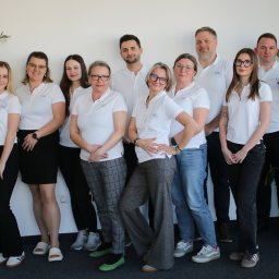 Zespół w białych koszulkach polo z logo firmy, stojący na tle białej ściany z roślinami doniczkowymi po bokach. Uśmiechnięte twarze, różnorodne stylizacje spodni.