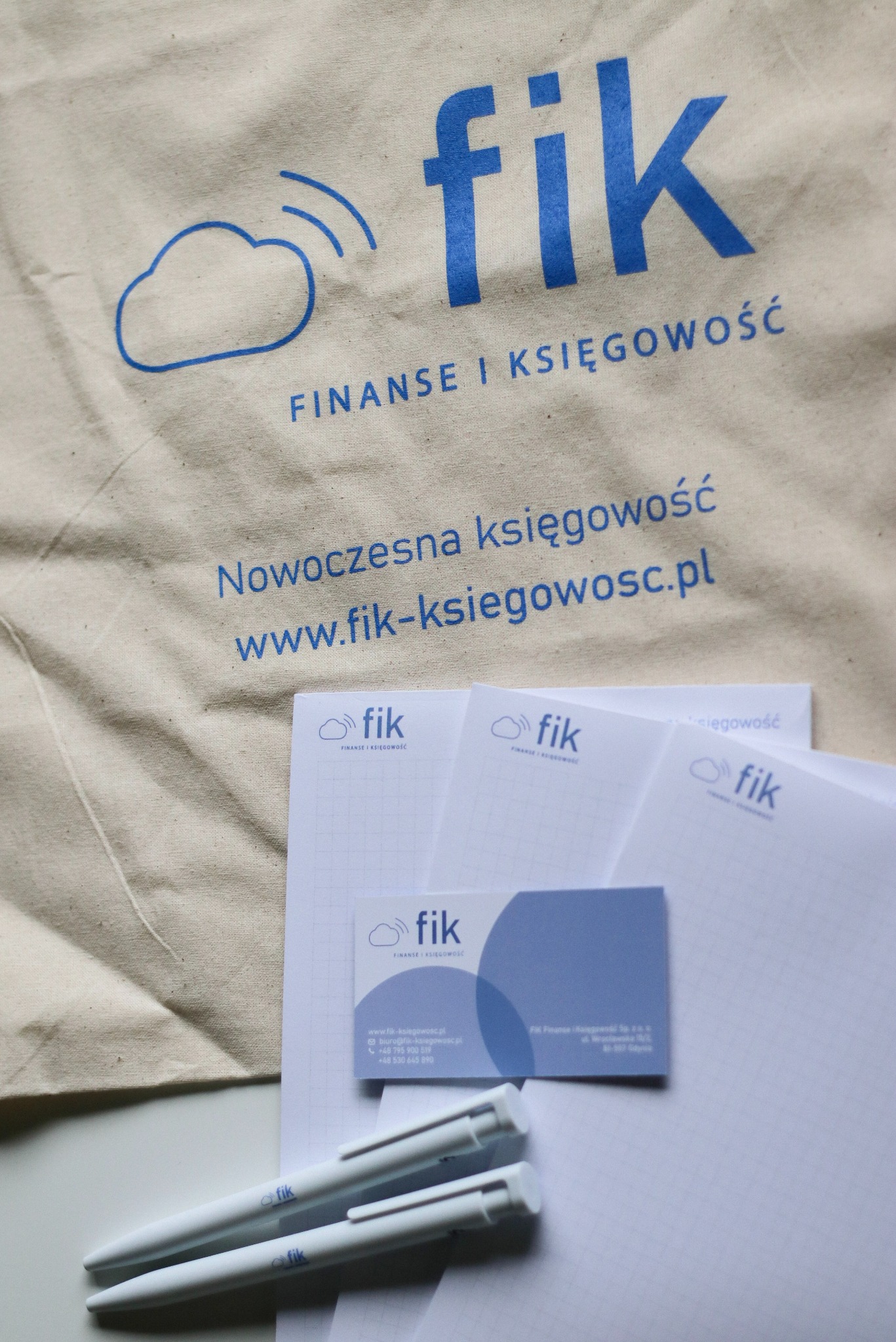 Torba bawełniana z logo 'fik Finanse i Księgowość' oraz wizytówki i długopisy z tym samym logo. Całość w tonacji błękitno-białej, na jasnym tle.