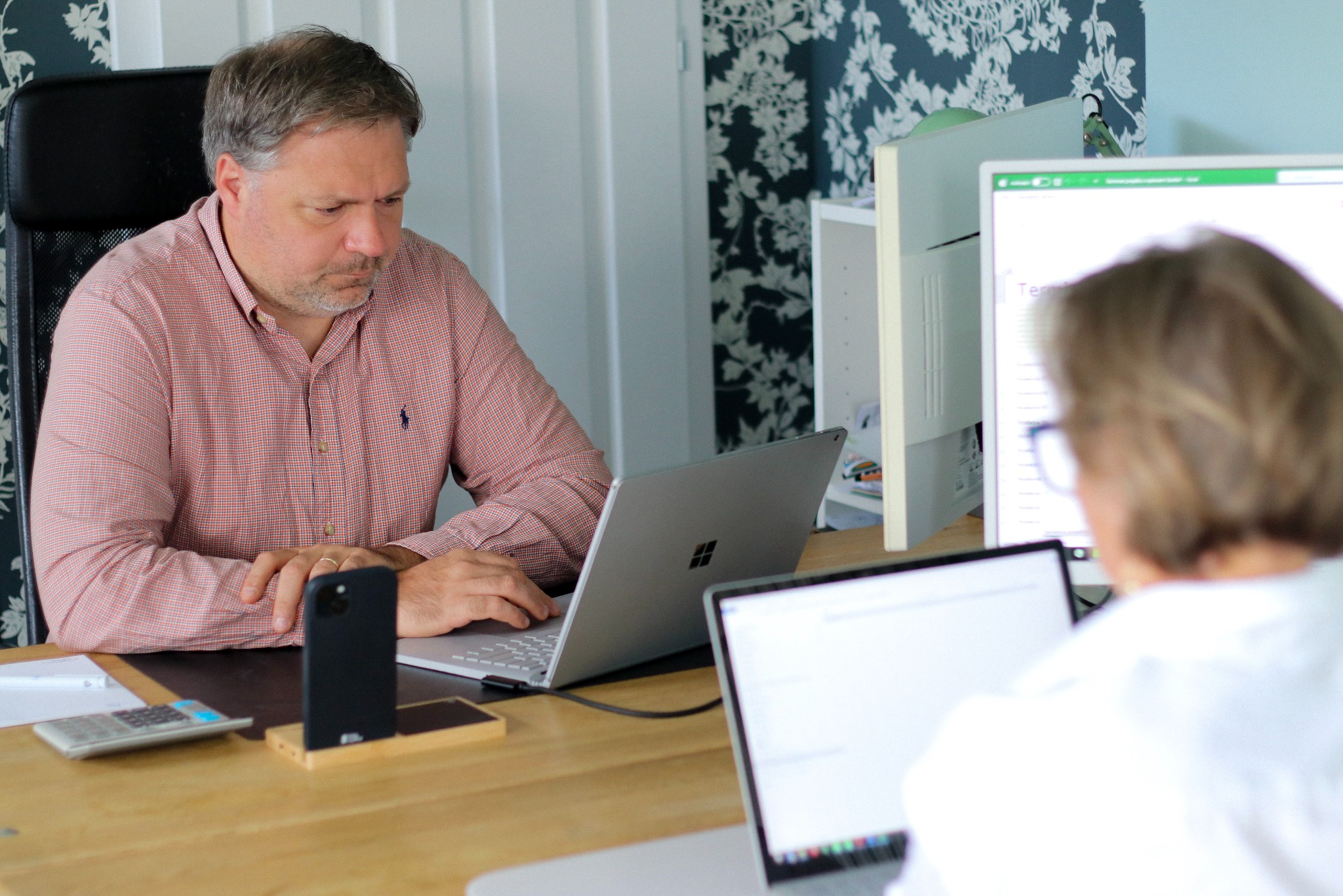 Mężczyzna w koszuli w kratę pracuje przy biurku z laptopem, telefonem i kalkulatorem. Drugi laptop i monitor w tle. Tapeta w kwiaty dodaje charakteru.